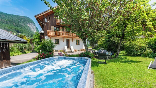 A hot tub in a lush garden with a wooden house and a mountain backdrop. - Detatched Chalet, 350m Samoens centre, 5 bedrooms, Jacuzzi/spa, pool table, fire (Samoëns)