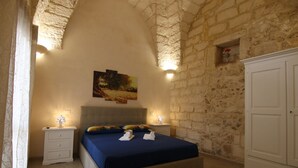 Een slaapkamer met een stenen muur, een bed met een blauwe spreideken, twee nachtkastjes met lampen, en een schilderij aan de muur.
