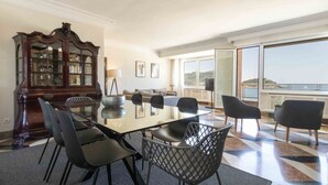 A spacious room with a large dining table, several chairs, a cabinet with glass doors, and a view of the sea through large windows.