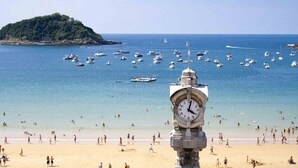 A beach with a clock tower in the foreground, boats in the water, and people on the beach.
