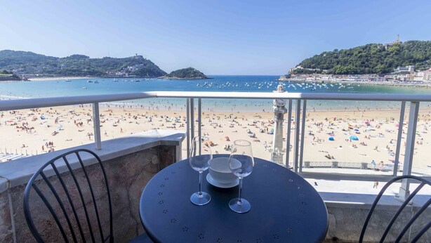 A balcony with a table, two wine glasses, and a view of a crowded beach and sea.