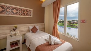 A hotel room with a bed, a nightstand with a phone and a lamp, a window with a view of a body of water and buildings, and a decorative wall panel. - Red Chilies Hotel (Surakarta)