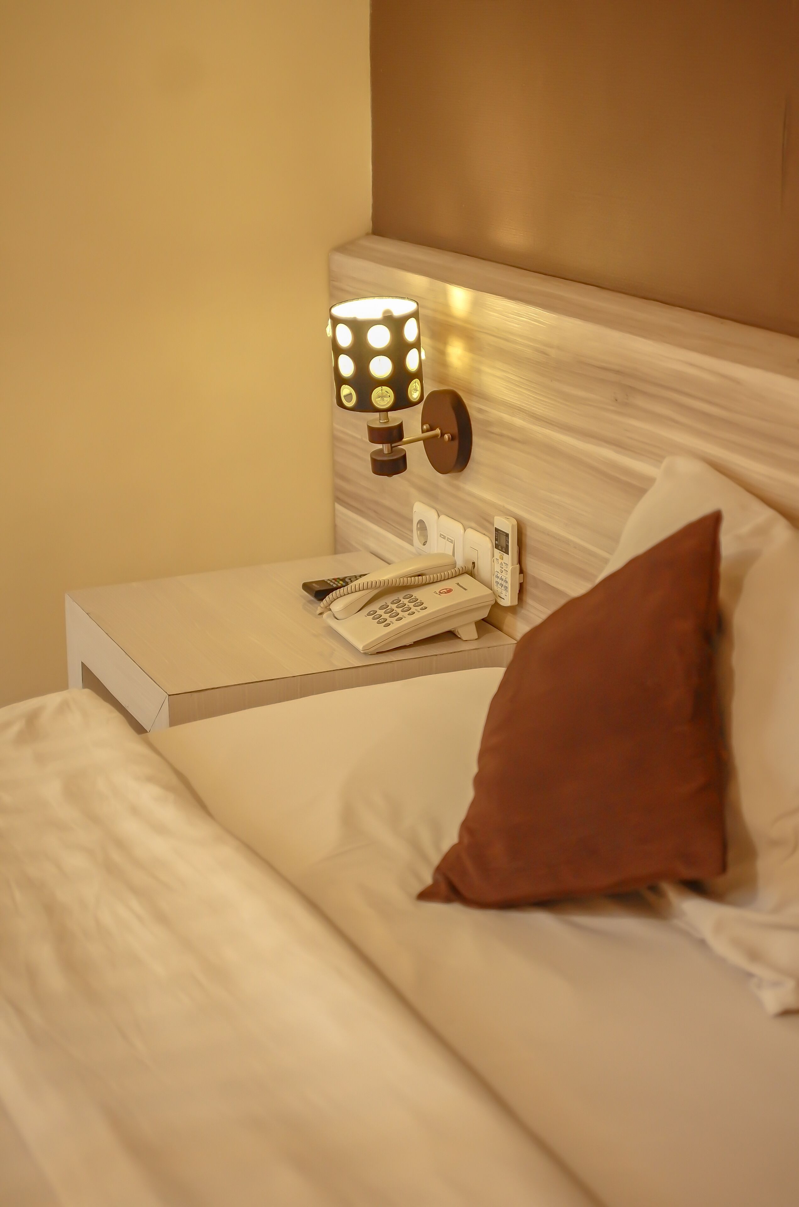 Una cama bien tendida con un cojín marrón, una lámpara de noche y un teléfono sobre una mesita de noche.