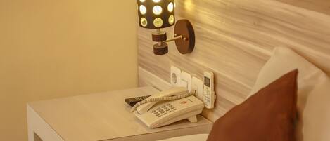 Una cama bien tendida con un cojín marrón, una lámpara de noche y un teléfono sobre una mesita de noche.