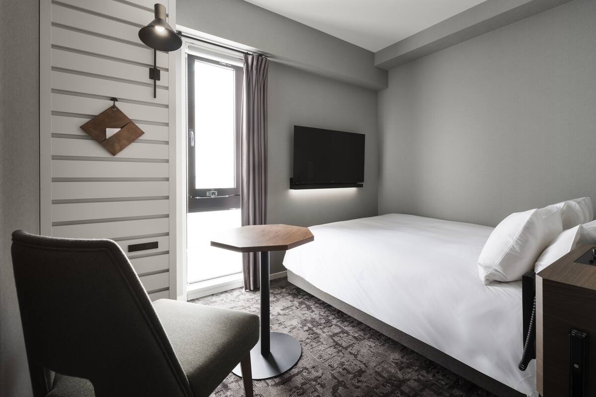 Ein Hotelzimmer mit einem Bett, einem Schreibtisch, einem Sessel, einem an der Wand befestigten Fernseher und einem Fenster mit Vorhängen.