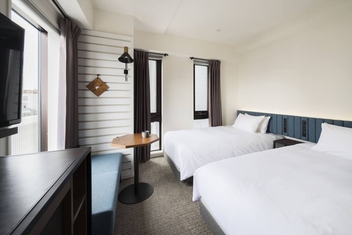 Ein Hotelzimmer mit zwei Betten, einem Schreibtisch, einem Stuhl, einem Fernseher und einem Fenster mit Vorhängen.