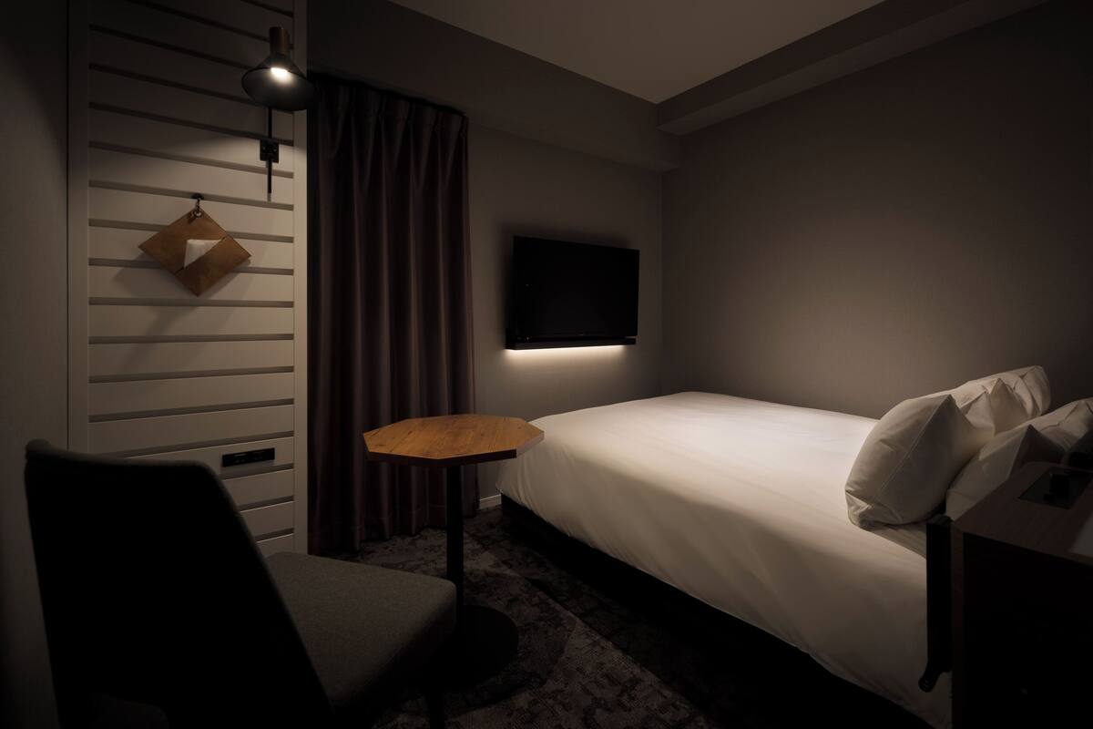 Ein Hotelzimmer mit Bett, einem kleinen Tisch, einem Sessel, einem an der Wand montierten Fernseher und einer Pendelleuchte.