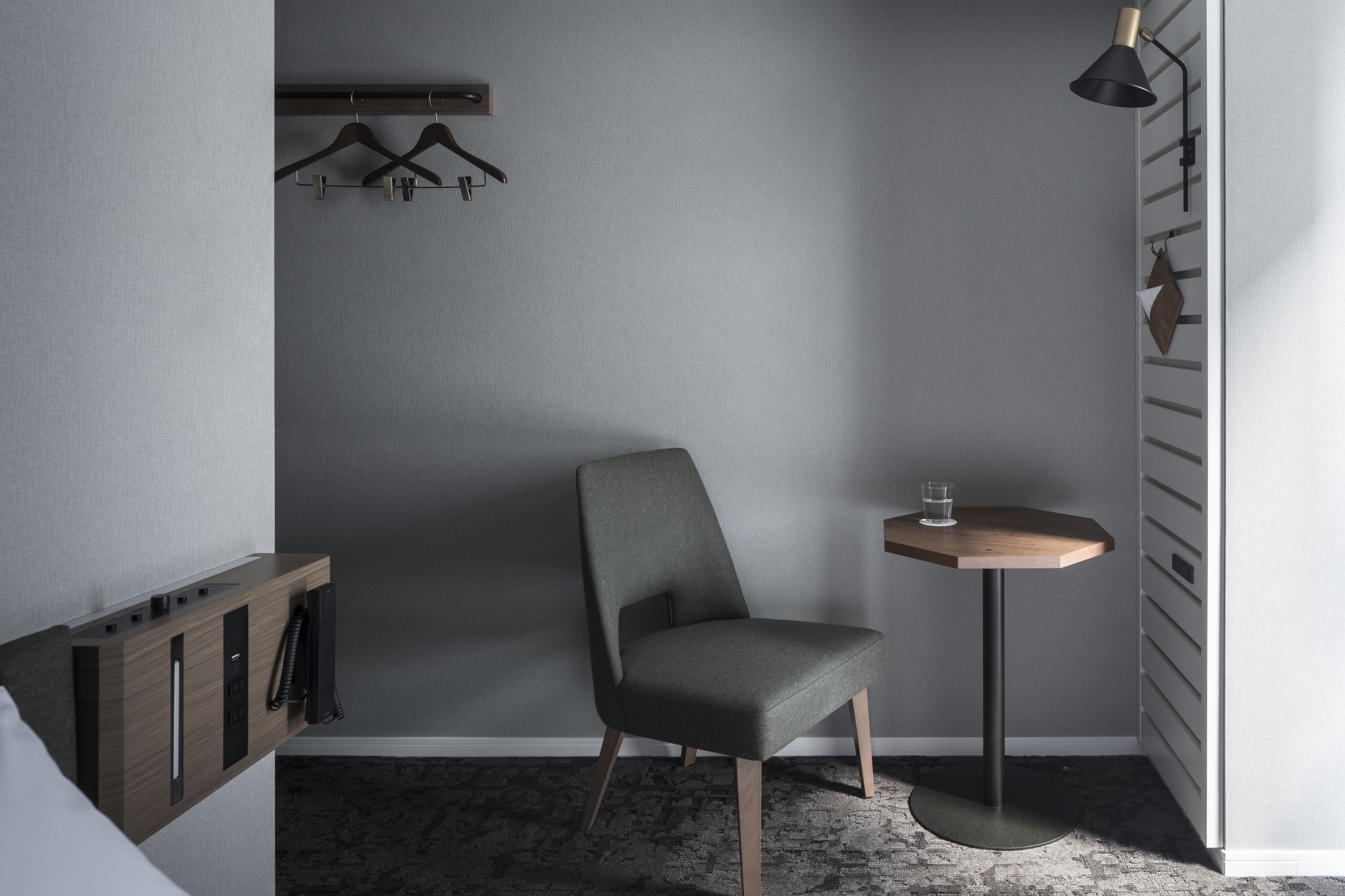 A modern room with a grey upholstered chair, a small round table with a glass, a wall-mounted coat rack, and a wooden cabinet with a telephone.