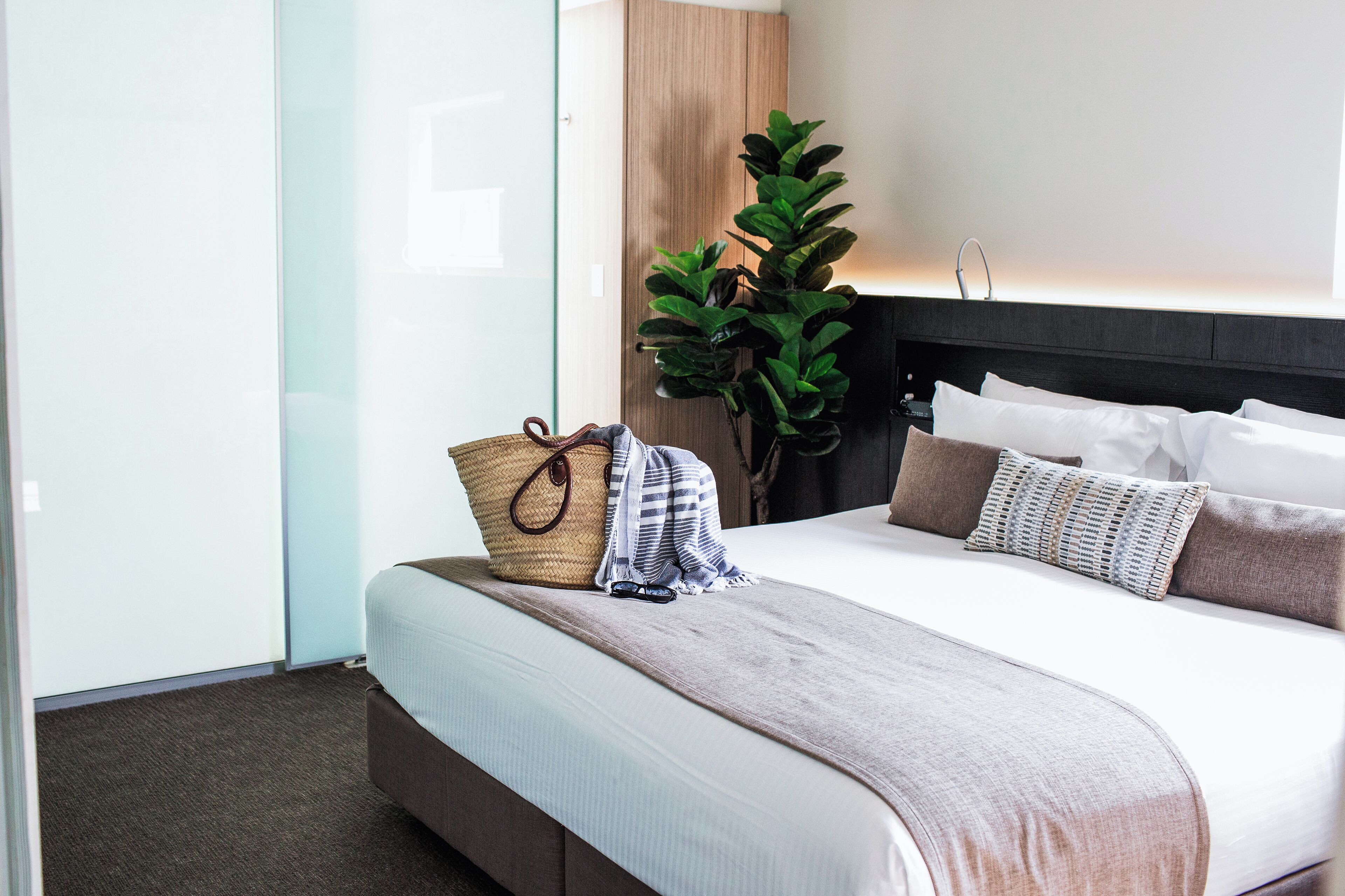 A neatly made bed with a headboard, a bedside basket, and a potted plant.