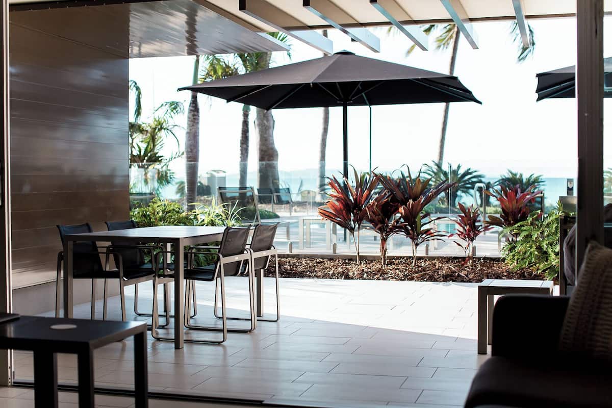 Een terras met een tafel, stoelen, een grote parasol en tropische planten.
