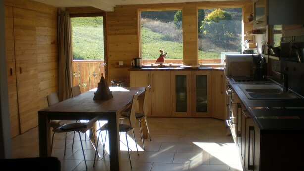 Una cabaña de madera con comedor, cocina y ventanas amplias que dan a un paisaje cubierto de hierba.