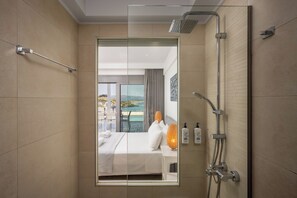 A modern bathroom with a glass shower enclosure, a visible bedroom with a bed and a view, and a bathroom counter with toiletries.