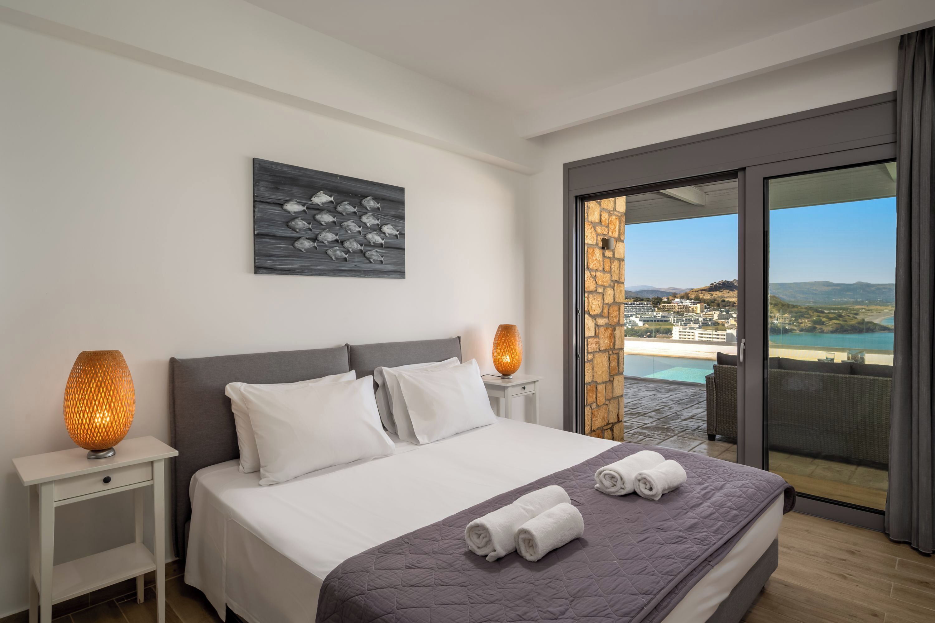 A hotel room with a bed, two bedside lamps, a painting, and a balcony with a view of the beach and buildings.
