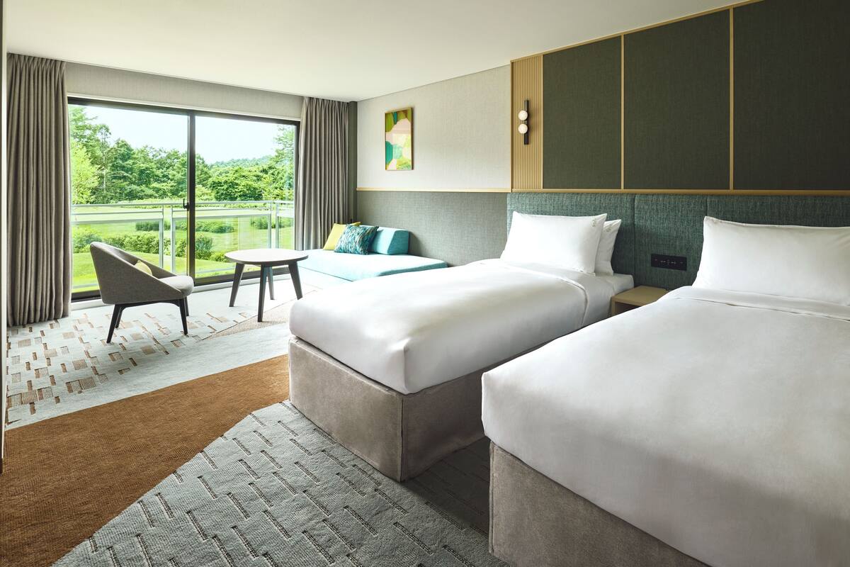A hotel room with two beds, a small table, a chair, and a view of greenery through large windows.