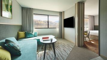 Une chambre d’hôtel avec un canapé, une petite table sur laquelle se trouvent un livre et une tasse, une télévision et une fenêtre donnant sur des arbres.
