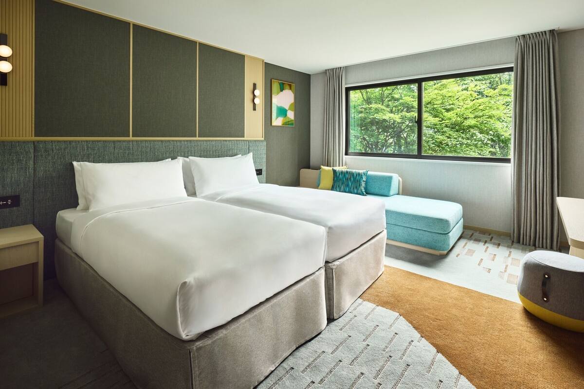 A hotel room with two beds, a sofa, a window with a view of trees, and a small table.