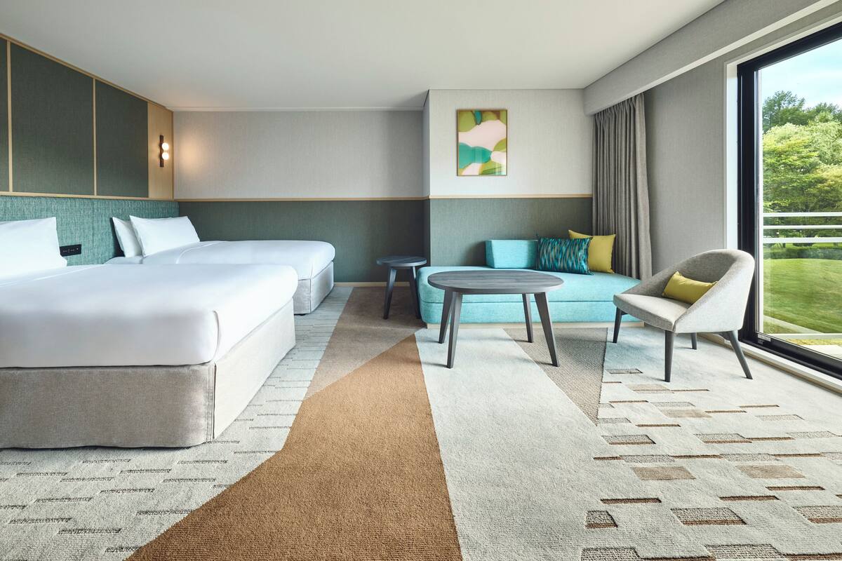 Habitación de hotel con dos camas, un sofá, una mesita pequeña y un ventanal grande que ofrece vistas a zonas verdes.