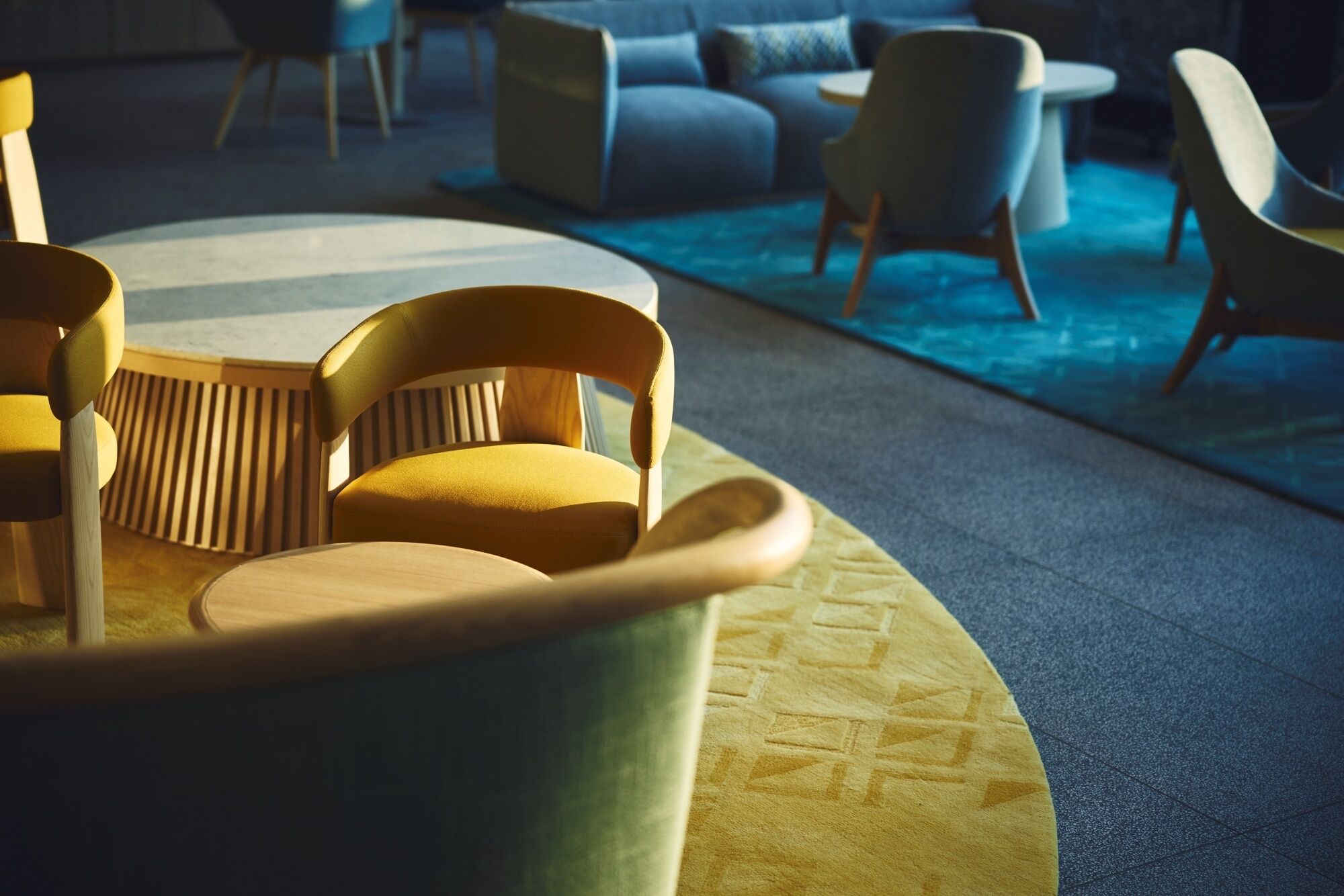 A modern lounge area with a round table and yellow chairs, a patterned rug, and a blue sofa in the background.