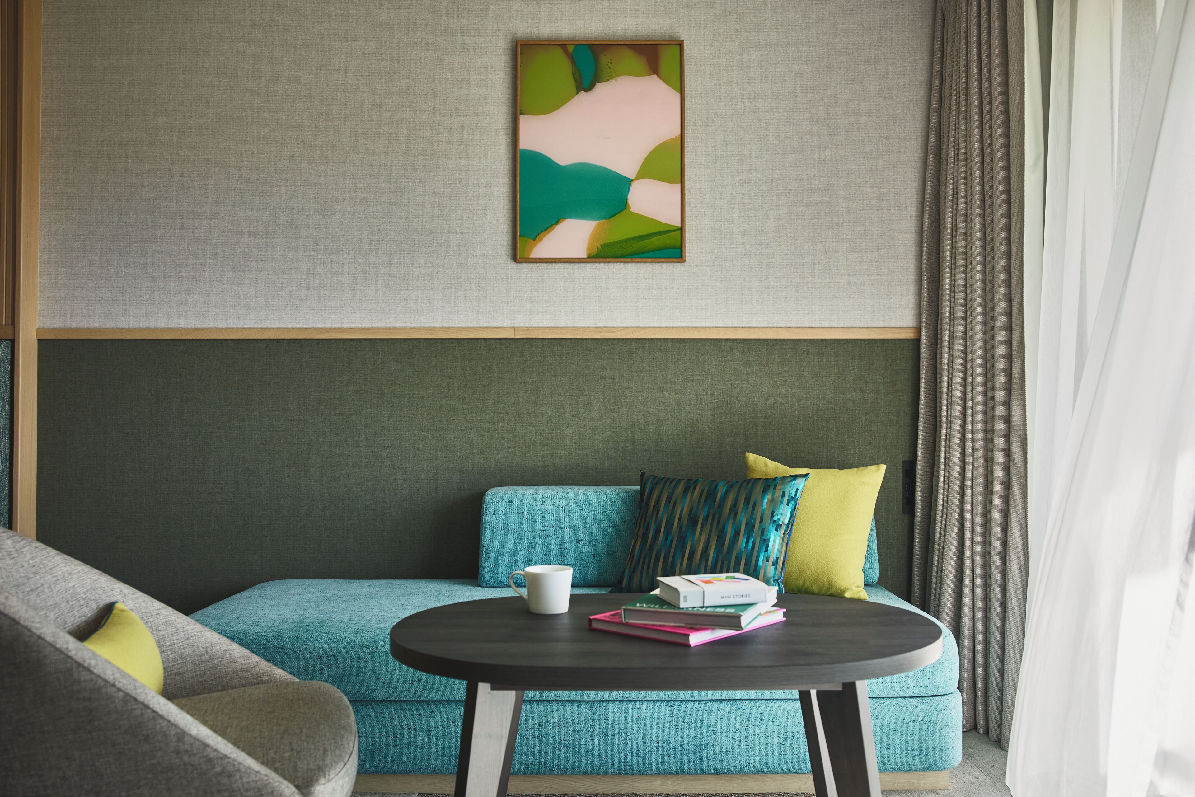 A modern room with a teal sofa, a round coffee table with books, and a framed abstract painting on the wall.