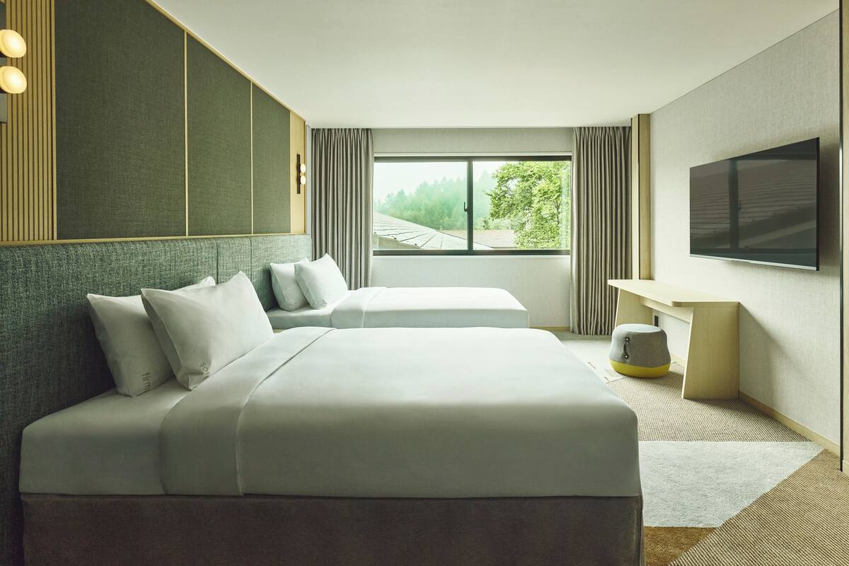 A modern hotel room with two beds, a flat-screen TV, and a large window offering a view of trees.