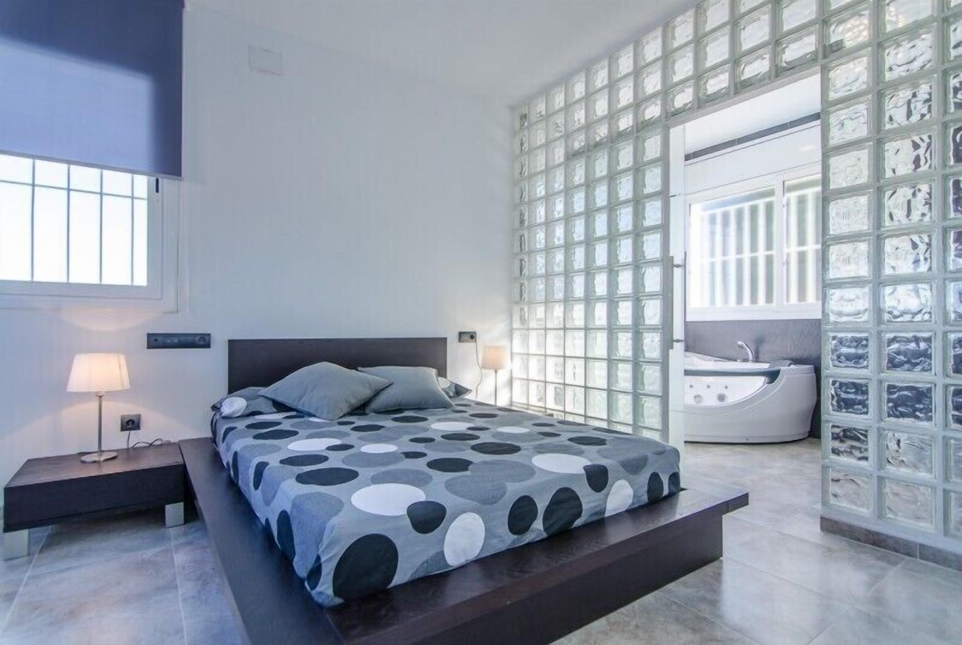 A modern bedroom with a bed, bedside tables, a large window, and a bathroom with a bathtub.