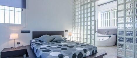A modern bedroom with a bed, bedside tables, a large window, and a bathroom with a bathtub.