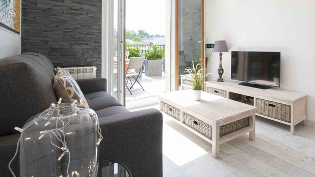 En moderne stue med sofa, sofabord i glas, fjernsyn på træfod og en balkon med udendørs møbler.