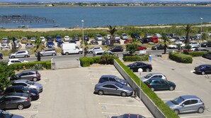 Un parking rempli de voitures et où se trouve un fourgon blanc, d’où l’on a vue sur une plage et sur la mer.