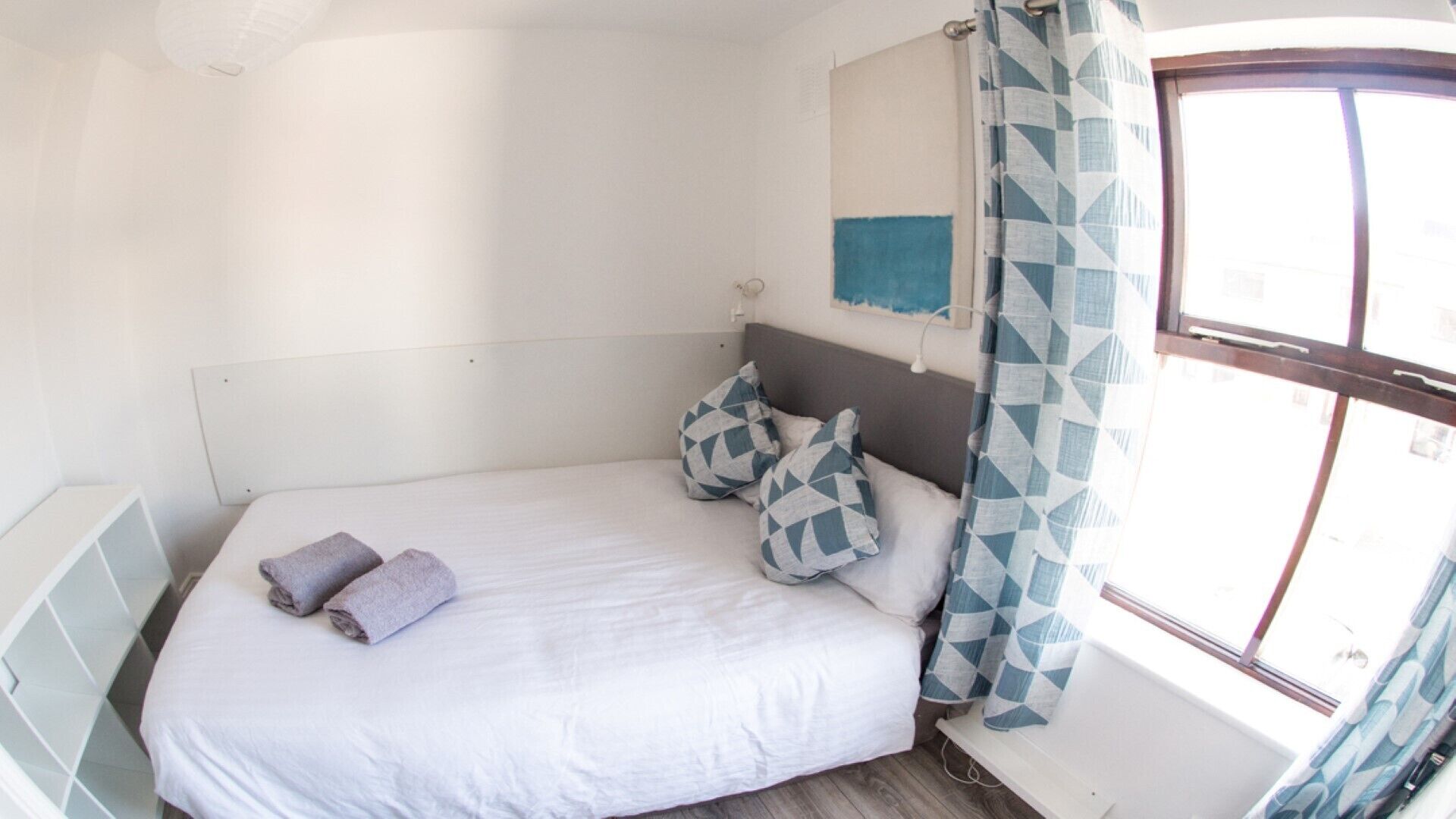 Een slaapkamer met een bed, kussens, een hoofdbord, een raam met gordijnen en een wandlamp.