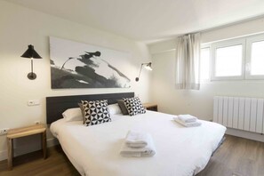 Una cama bien hecha con sábanas blancas y toallas dobladas, un cuadro abstracto grande en la pared, dos lámparas de noche y una ventana con cortinas.