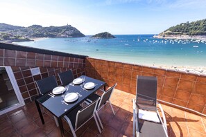 Una terraza con mesa lista para comer, con vista a una playa con barcos y un paisaje montañoso.