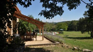 Un porche cubierto con mobiliario exterior, una pérgola y vistas a un paisaje verde con árboles y una piscina.