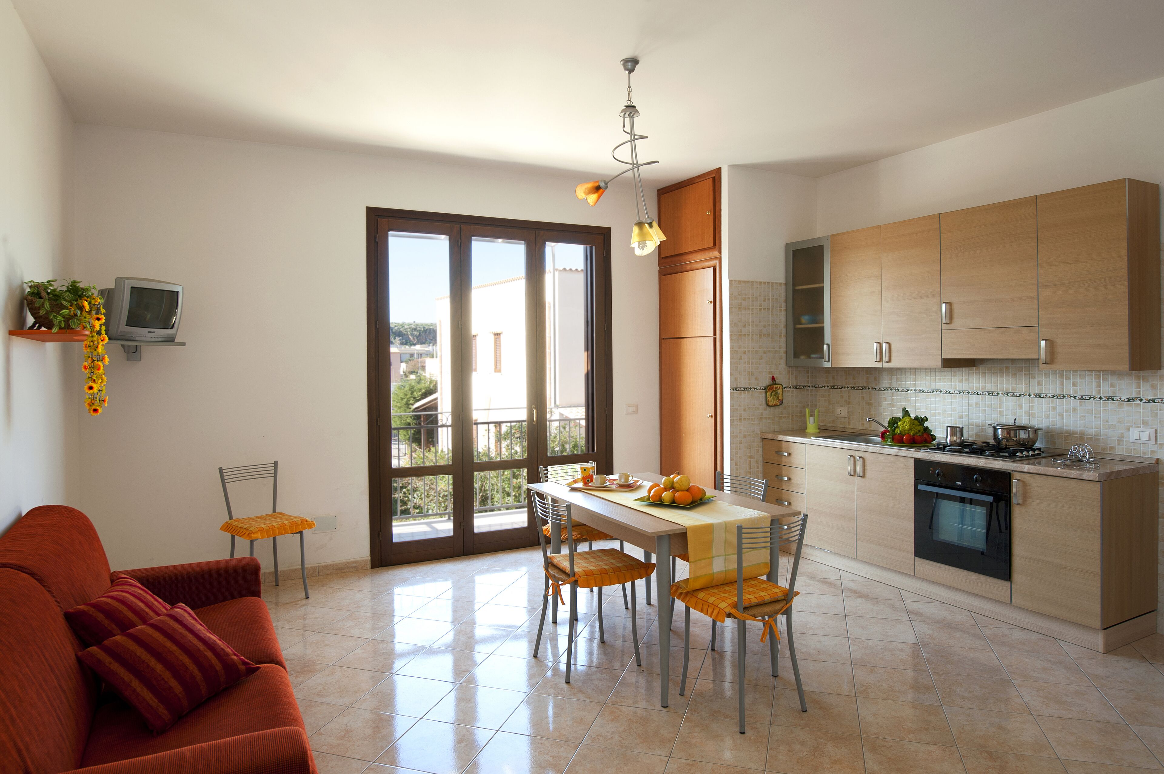 A modern kitchen with a dining area, featuring a large window, a sofa, and a TV on a shelf.