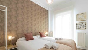 A hotel room with two beds, a patterned wallpaper, a bedside table with a lamp, and a window with curtains. - Urbieta | The Rentals Collection (San Sebastián)