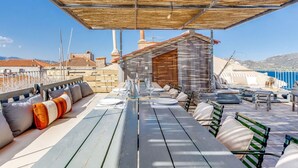 An outdoor seating area with wooden tables and benches, a covered section with a bamboo roof, and a view of the sea and buildings in the background. - Cosy apartment for 4 people with A/C, WIFI, TV and terrace (Porto-vecchio)