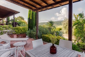 An outdoor patio with wicker furniture, a wooden table, and a view of a lush garden and hills.