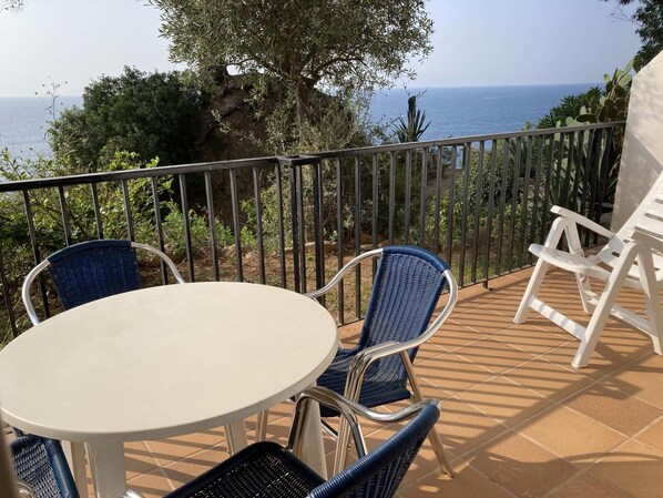 A terrace with a round table and blue and white wicker chairs, overlooking a rocky coastline and the sea.