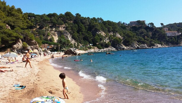 Ein Strand mit klarem, blauem Wasser, Menschen beim Schwimmen und ein Kind, das am Sand entlanggeht.