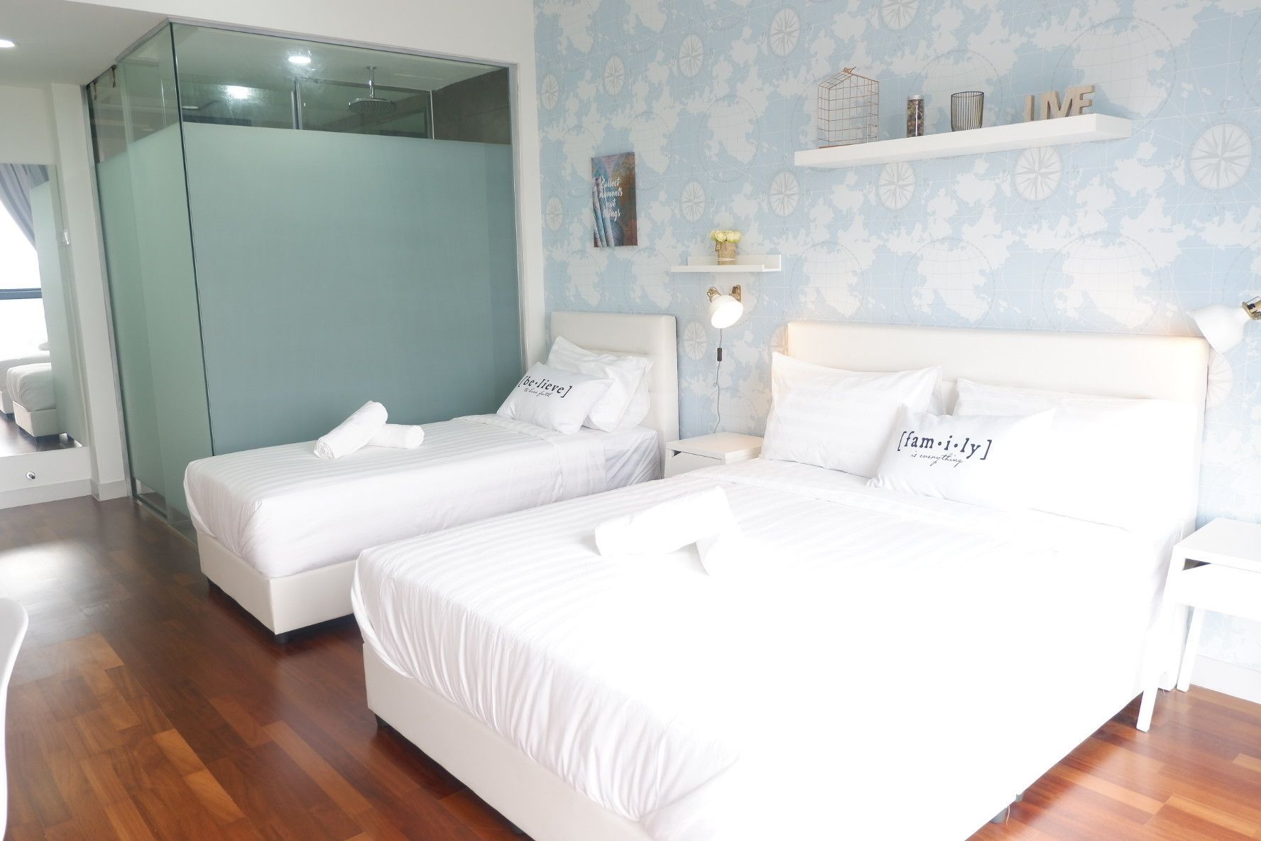 A hotel room with two beds, a glass partition, a world map wallpaper, and a shelf with decorative items.
