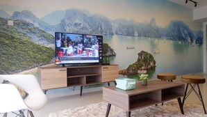 A living room with a large TV displaying a news channel, a wooden entertainment unit, a white chair, a fluffy rug, and a view of a coastal landscape through the window. - Bora Residence (Johor Bahru)