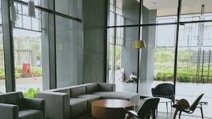 A modern lobby with a grey sectional sofa, a round coffee table, and two chairs. Large windows offer views of greenery outside. - Bora Residence (Johor Bahru)