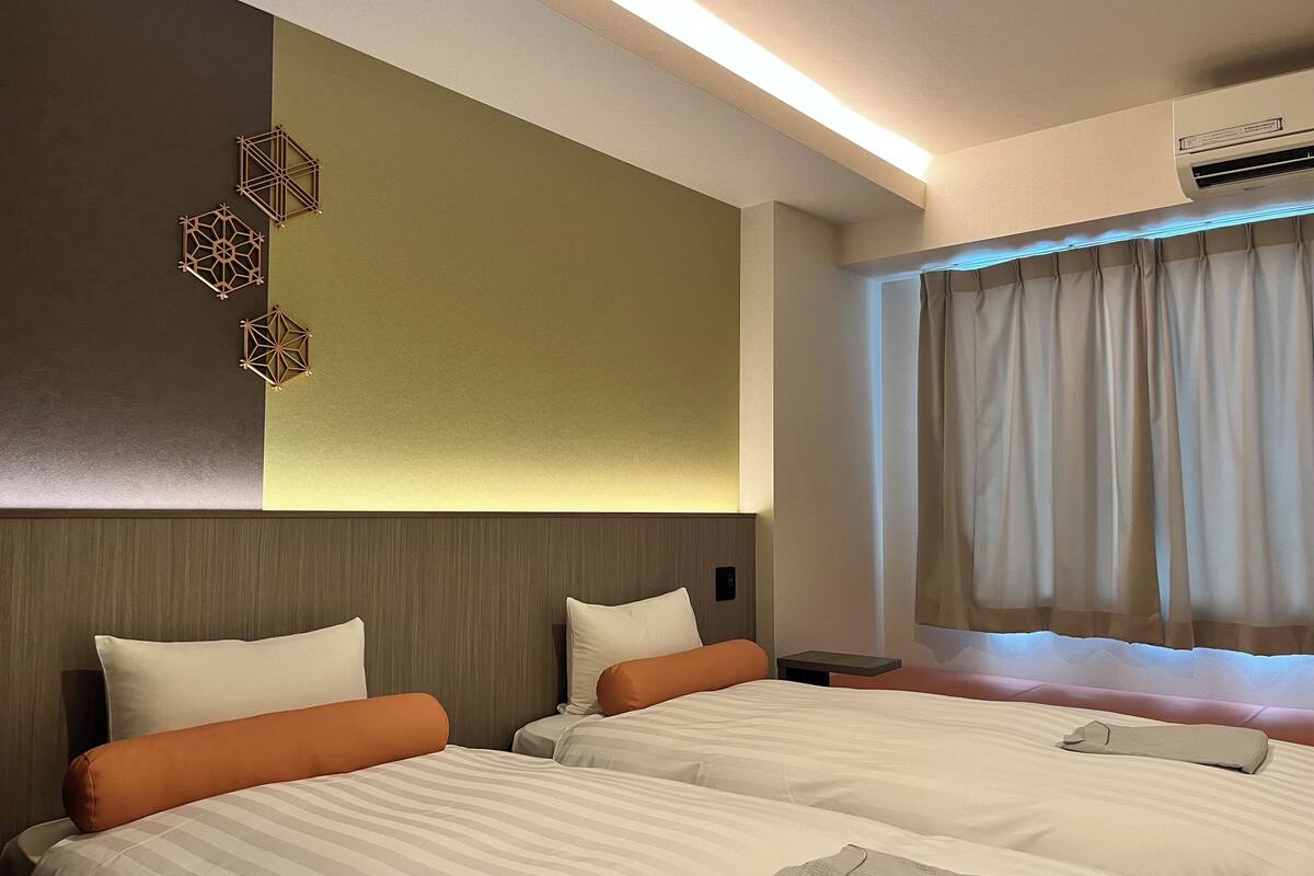 A hotel room with two beds, a nightstand, an air conditioning unit, and a window with curtains.