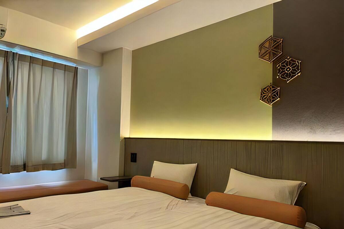 A hotel room with a large bed, two pillows, a bedside table, a window with curtains, and a wall with geometric wall art.
