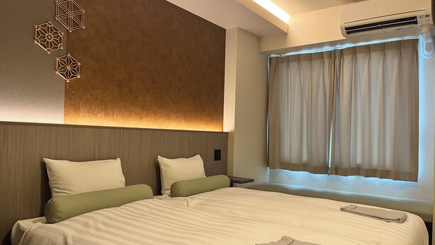 A hotel room with a large bed, two pillows, a bedside table, a wall-mounted air conditioner, and a window with curtains. - Hotel Japanesque Kyoto Station ZEQUU (Kyoto)