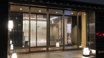 A hotel entrance with a modern design, featuring a glass door with vertical blinds, a reception desk, and a well-lit interior.