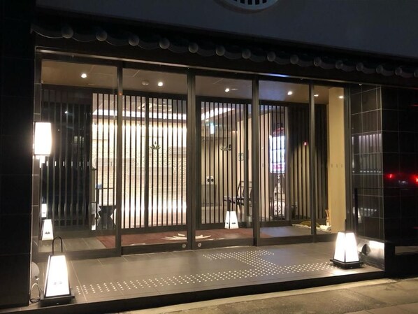 A hotel entrance with a modern design, featuring a glass door with vertical blinds, a reception desk, and a well-lit interior.