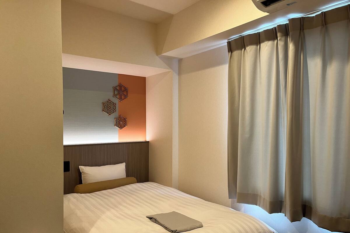 A hotel room with a bed, a bedside table, a wall-mounted air conditioner, and a window with curtains.