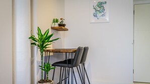 A corner room with a small wooden table, two black chairs, a potted plant, and a wall-mounted shelf with decorative items.