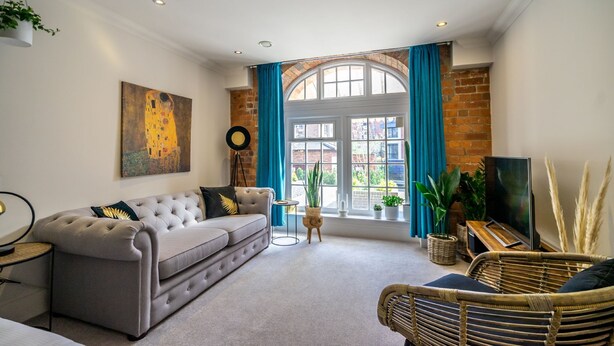 A living room with a grey sofa, a wicker chair, a TV, and a large window with blue curtains.
