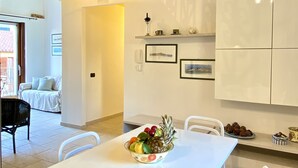 A dining area with a white table and chairs, a fruit bowl on the table, and a kitchen area with cabinets and a light fixture.